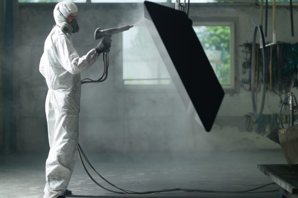 view of a worker wearing a full white protective suit and breathing mask, sand blasting a metal crate hung from a metal beam in the ceiling of an industrial hall