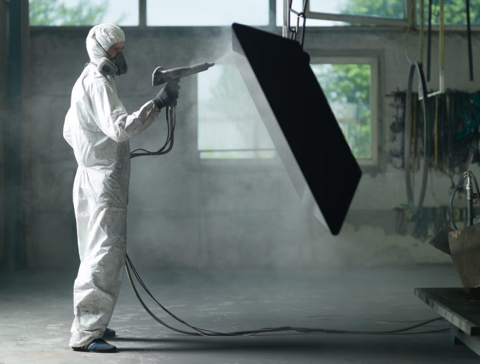 view of a worker wearing a full white protective suit and breathing mask, sand blasting a metal crate hung from a metal beam in the ceiling of an industrial hall