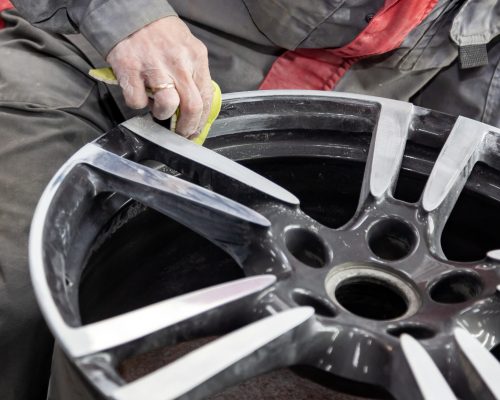 Master body repair man is working on preparing the surface of the aluminum wheel of the car for subsequent painting in the workshop, cleaning and leveling the disk with the help of abrasive material