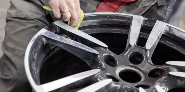 Master body repair man is working on preparing the surface of the aluminum wheel of the car for subsequent painting in the workshop, cleaning and leveling the disk with the help of abrasive material